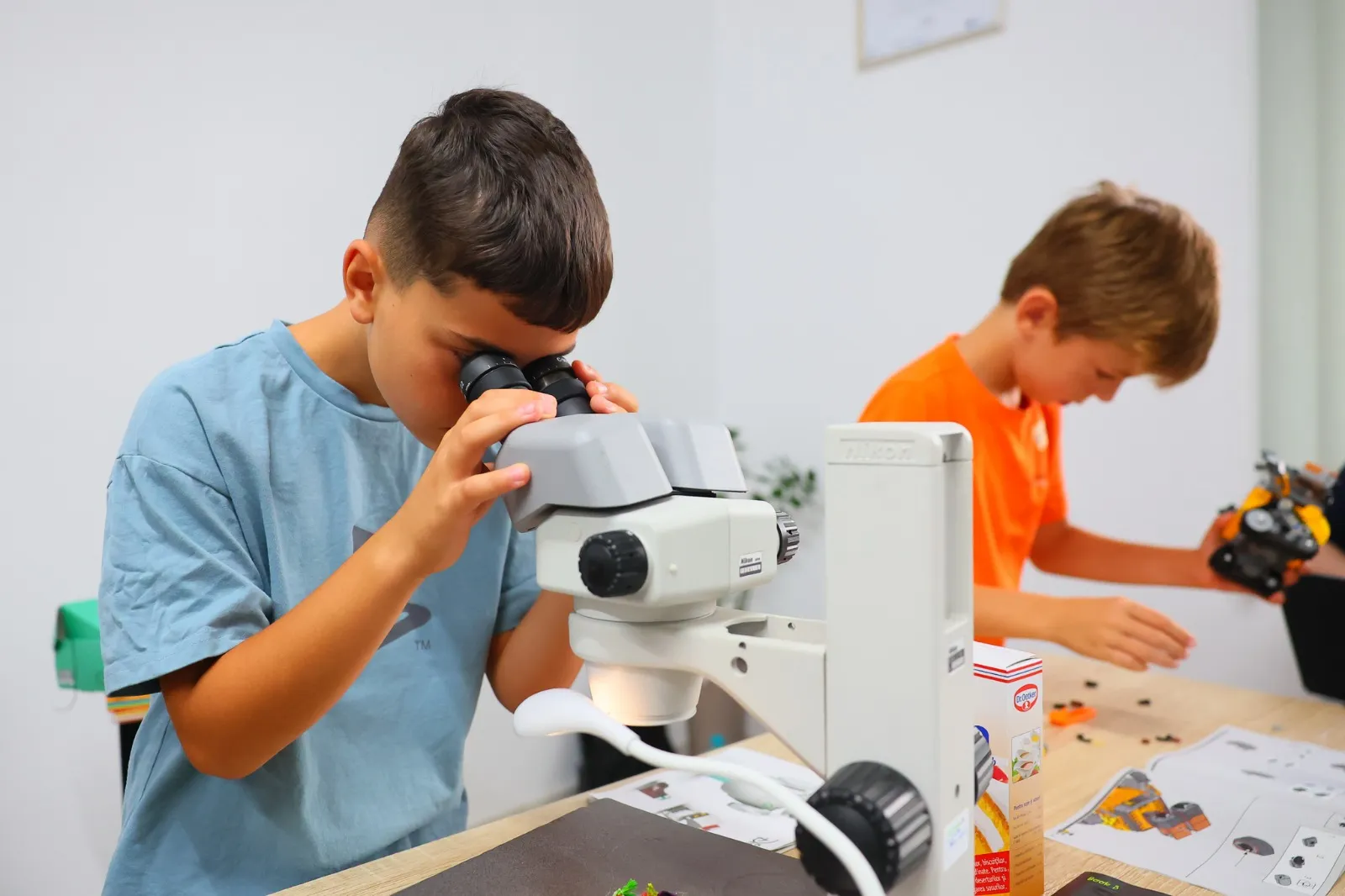 Băiat la stereomicroscop, cu un coleg care construiește un robot în fundal